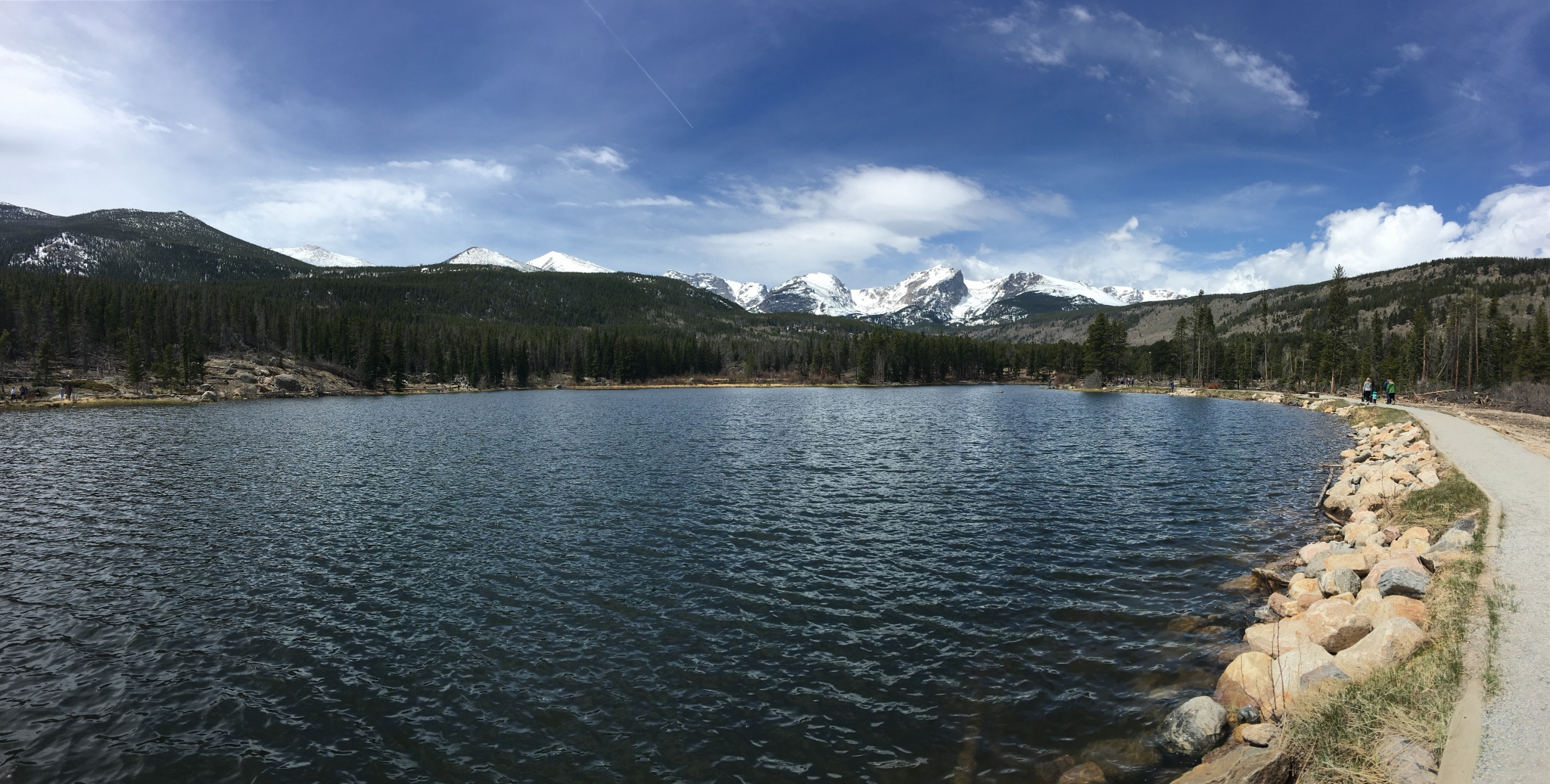 View of calm lake with pine forests and snow capped peaks in the backgroun on a sunny day